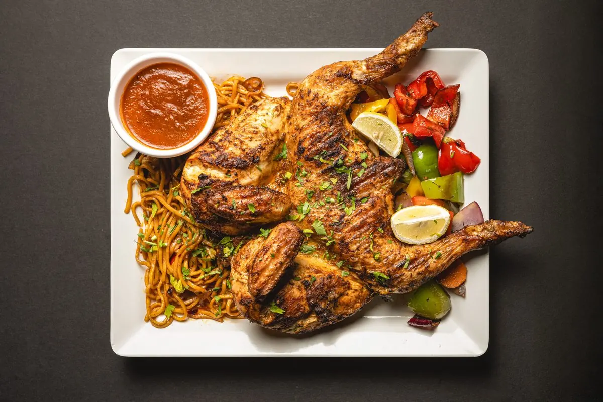A grilled chicken on noodles with peppers, lemon and a pot of dip